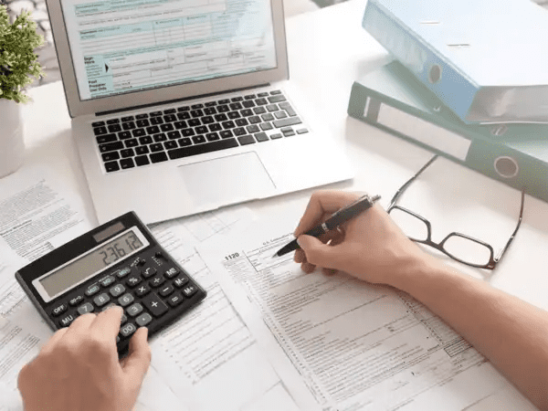 Person using a calculator and pen to fill out tax documents at a desk with a laptop and binders.