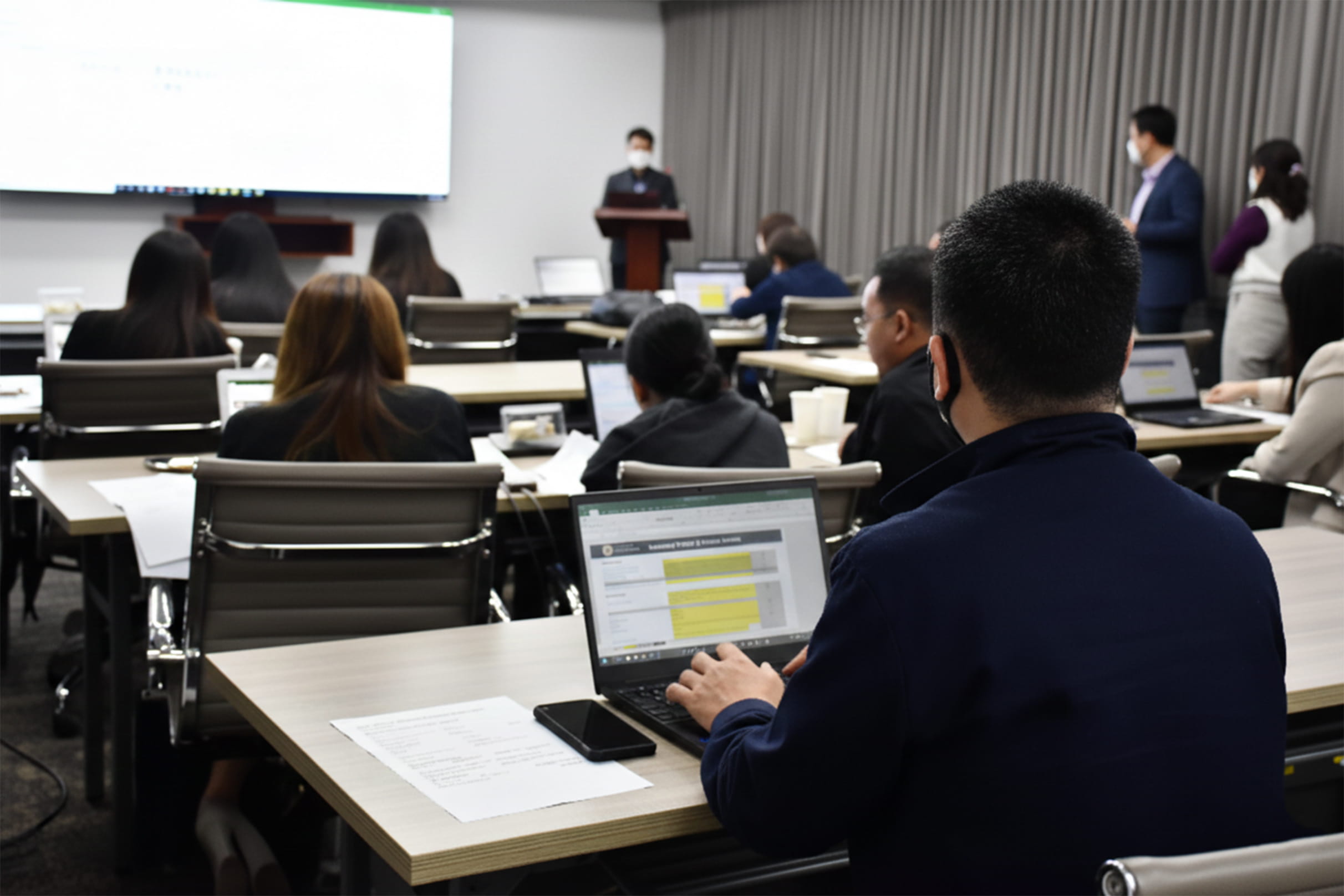 Attendees working on laptops while listening to a speaker presenting at a podium in a conference room setting.